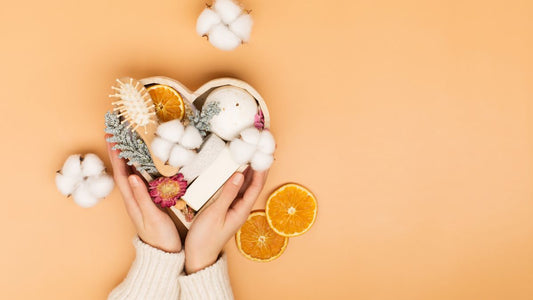 Hands holding a heart shape basket filled with self care items: a brush, cotton balls, oranges, etc